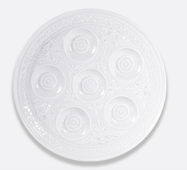 The Bernardaud Louvre Condiment Tray is a round, white, ornate plate with seven circular indentations and intricate patterns, inspired by classic Limoges France craftsmanship.