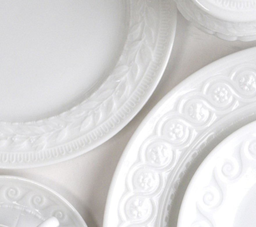 Close-up of several Bernardaud Louvre Condiment Trays, featuring ornate embossed rims inspired by French architecture, arranged overlapping on a light surface.