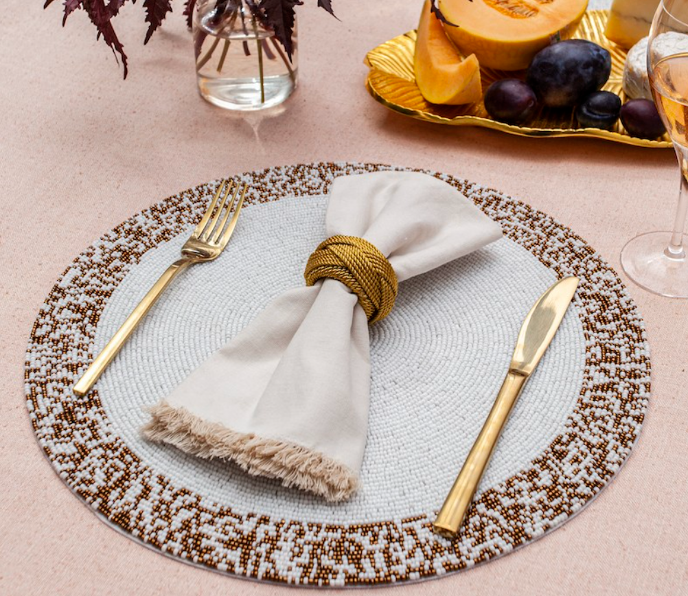The Von Gern Home Splatter Beaded Placemat Set of 4 adds elegance to your table, paired with a beige napkin, gold napkin ring, gold flatware, and complemented by a plate of fruit and a wine glass on a light tablecloth.