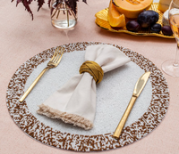 The Von Gern Home Splatter Beaded Placemat Set of 4 adds elegance to your table, paired with a beige napkin, gold napkin ring, gold flatware, and complemented by a plate of fruit and a wine glass on a light tablecloth.