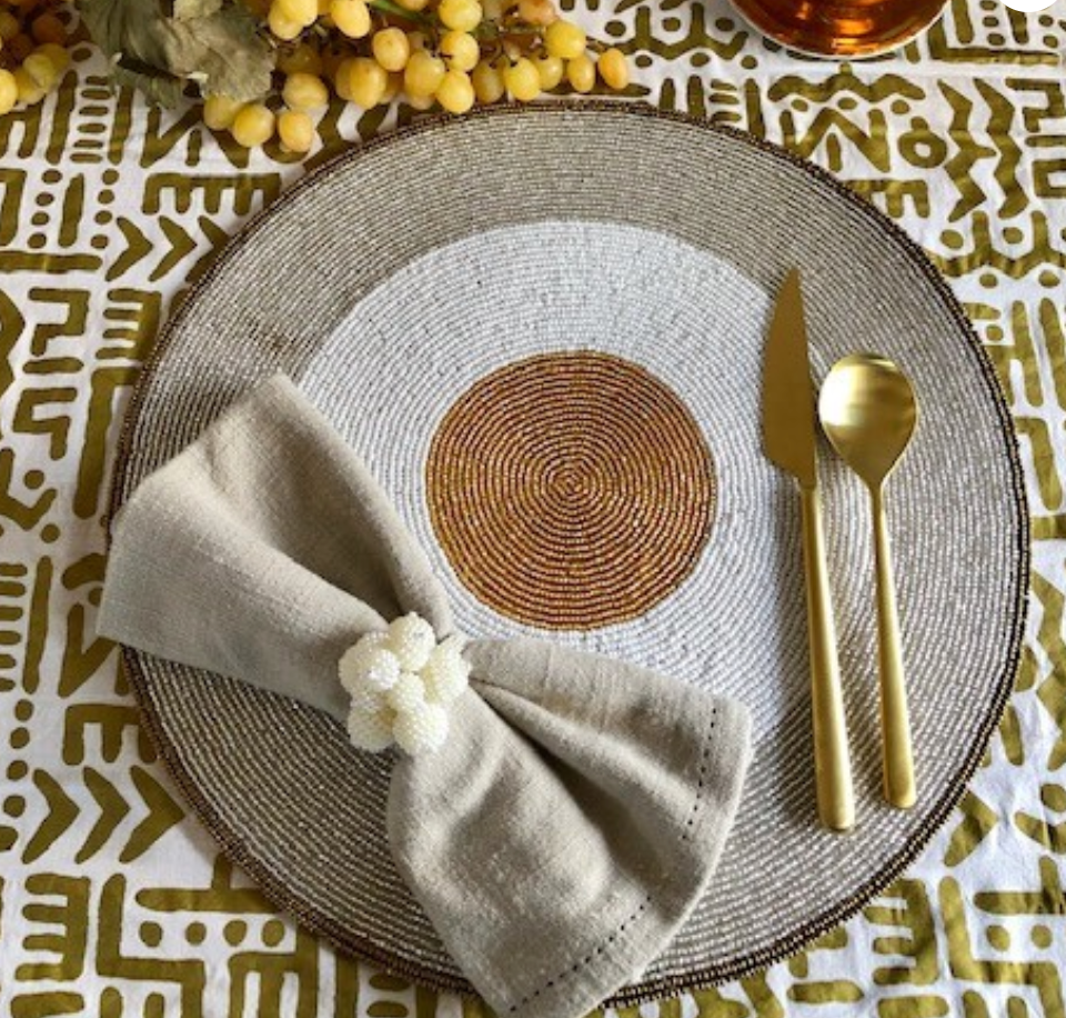 A sophisticated table decor is created with Von Gern Home’s Graphic Sparkle Placemats Set of 4, a folded beige napkin with a beaded napkin ring, gold-colored cutlery, and grapes in the background on a patterned tablecloth.
