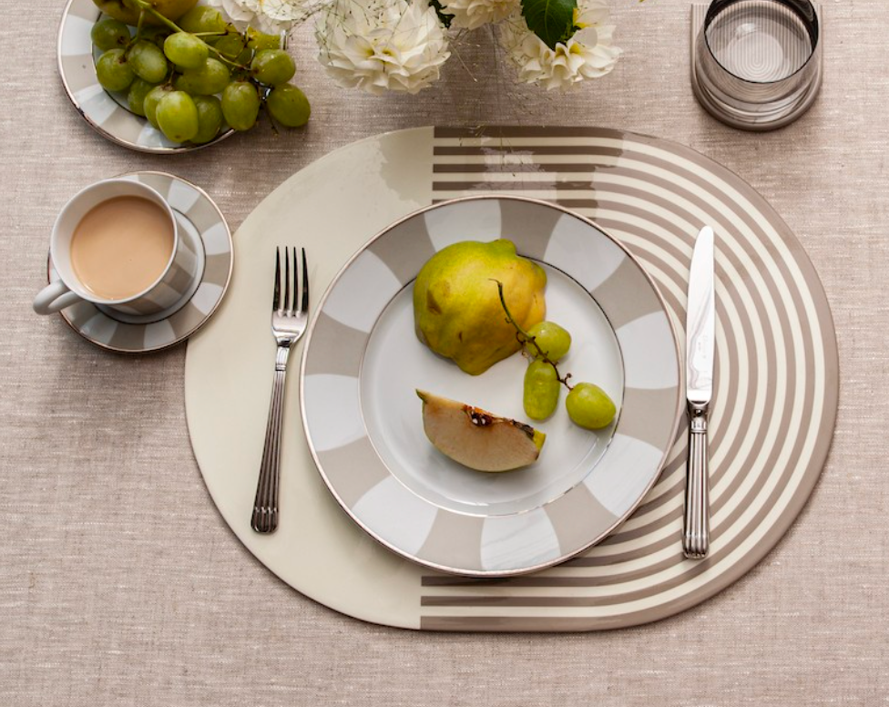 A modern table is set with fruit on a plate, coffee, flowers, and water atop Von Gern Home’s Stripes & Solids Lacquer Placemat Set 4 in beige and white.