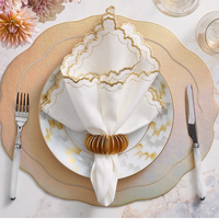 An elegant table setting features a gold-rimmed plate, gold charger, white and gold napkin in a Kim Seybert Demilune Napkin Ring (Gold Set of 4), silverware on each side, wine glasses, and pink flowers in the upper left corner.