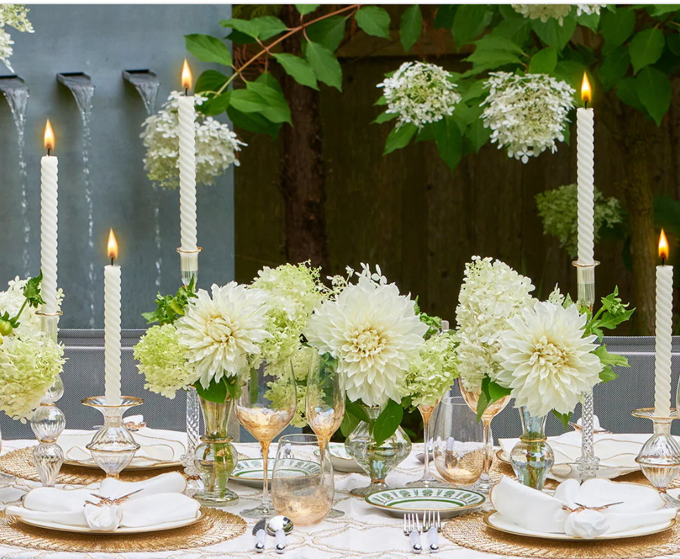 An elegant outdoor table set with white flowers, tall lit candles in Kim Seybert Clear Rib Gold Detail Trumpet Vases, gold-rimmed glassware, white napkins, and a white tablecloth. Lush greenery and blooming white hydrangeas provide a stunning background.