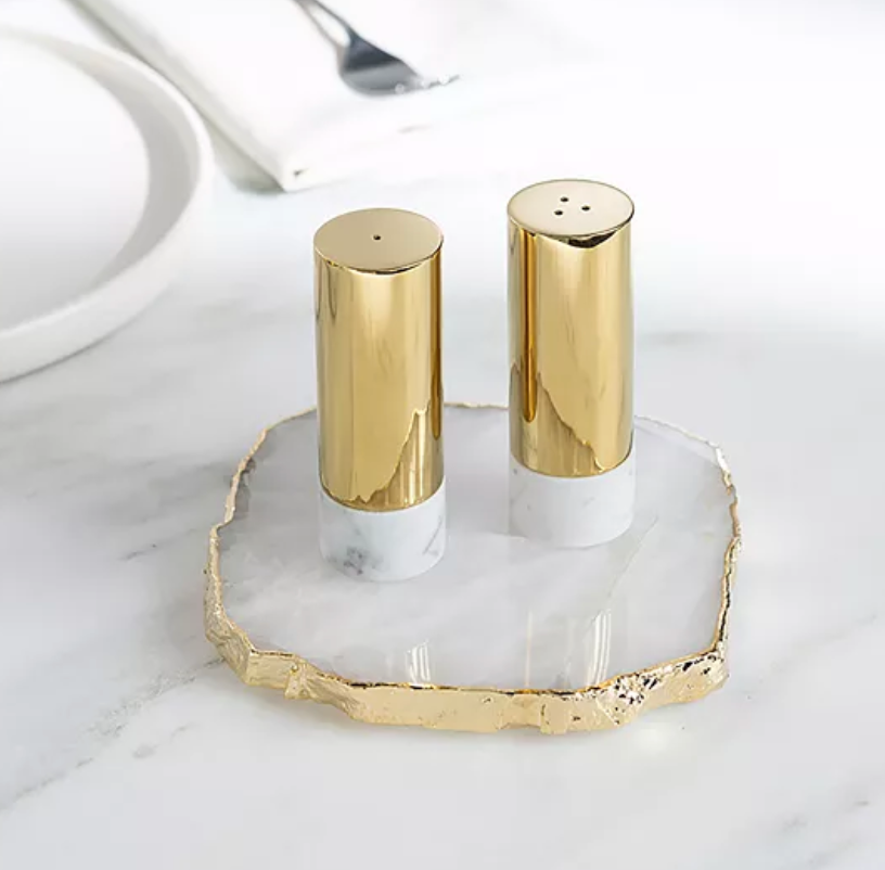 The Anna New York Dual Salt & Pepper Shakers Marble & Gold Set of 2 features elegant gold and white shakers on an Italian Carrara marble tray, displayed on a white surface with a plate, napkin, and fork in the background.
