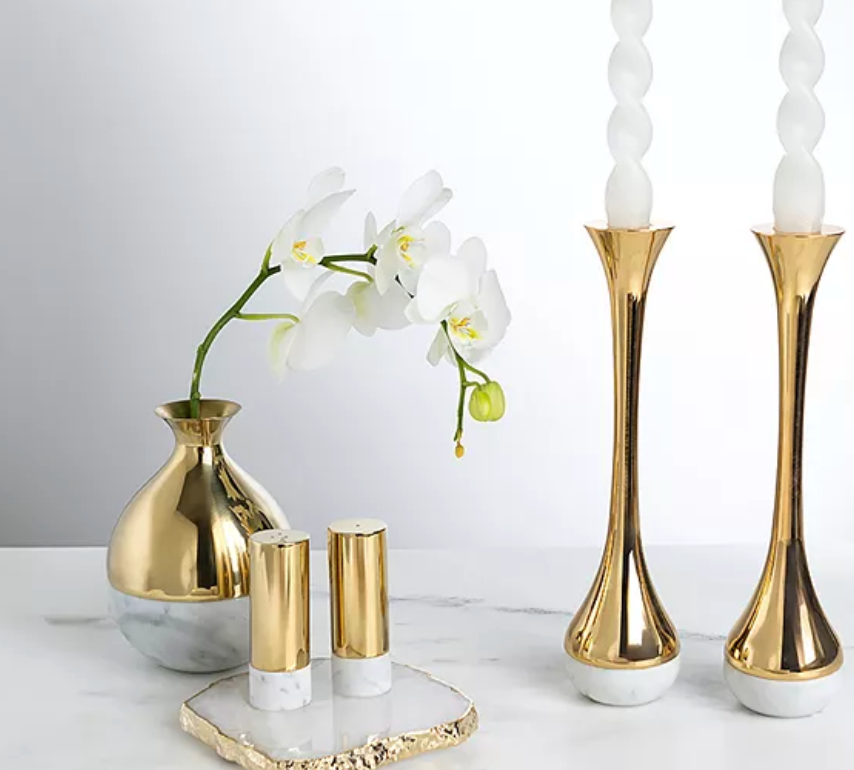 A gold and marble vase with white orchids sits next to two gold and white candlesticks and the Anna New York Dual Salt & Pepper Shakers Marble & Gold Set of 2 on a sleek Italian Carrara Marble surface.