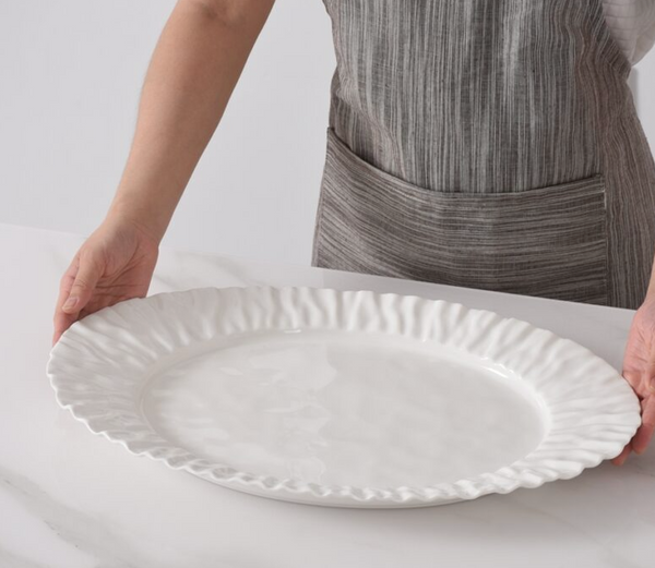 Wearing a striped gray apron, someone places the PAMPA BAY Mascali Bianca Serving Platter—microwave safe porcelain with wavy edges—on a white marble countertop.