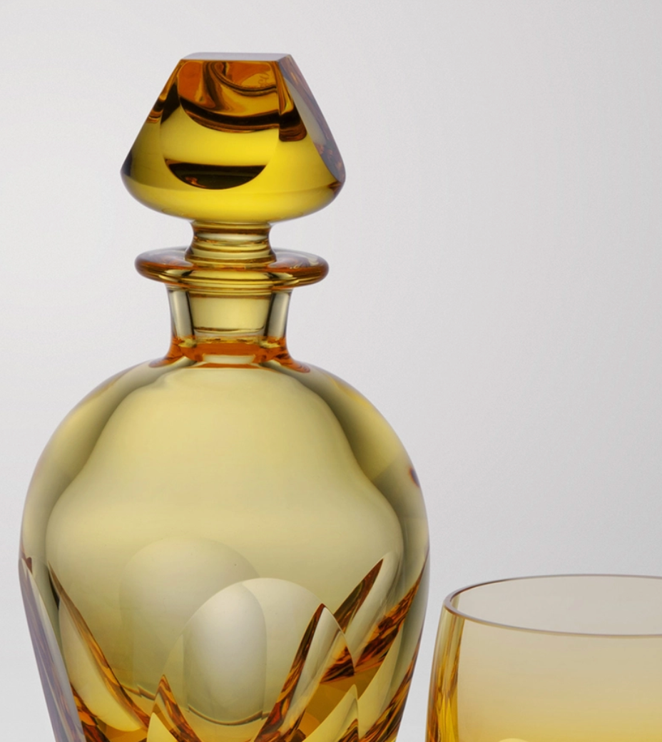 A close-up of the Moser Bar Decanter 750 ml, an amber crystal carafe with a faceted stopper beside a matching glass, both elegantly displayed against a plain light background.