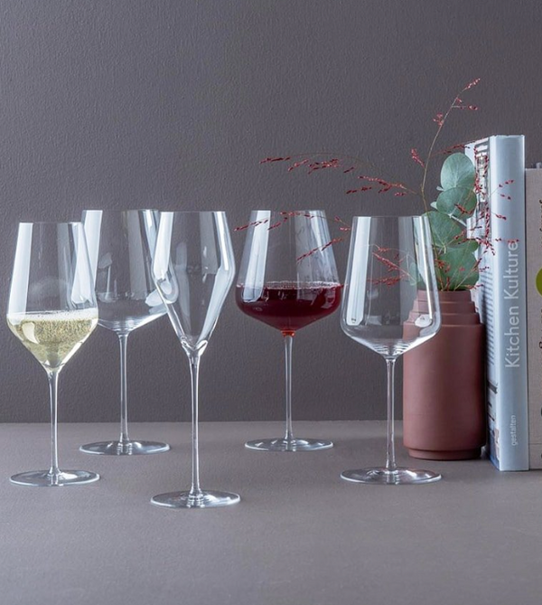 Six elegant, lead-free wine glasses—including a Zalto Bordeaux Glass by Grand Cru Selections/Zalto—stand on a grey surface before a dark grey wall, with two filled with white and red wine near a vase and books in the background.