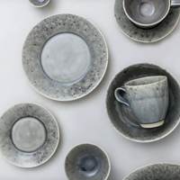Costa Nova’s Madeira Grey Dinnerware features stoneware plates, bowls, and cups in mottled grey and white finishes, artistically arranged on a light surface.
