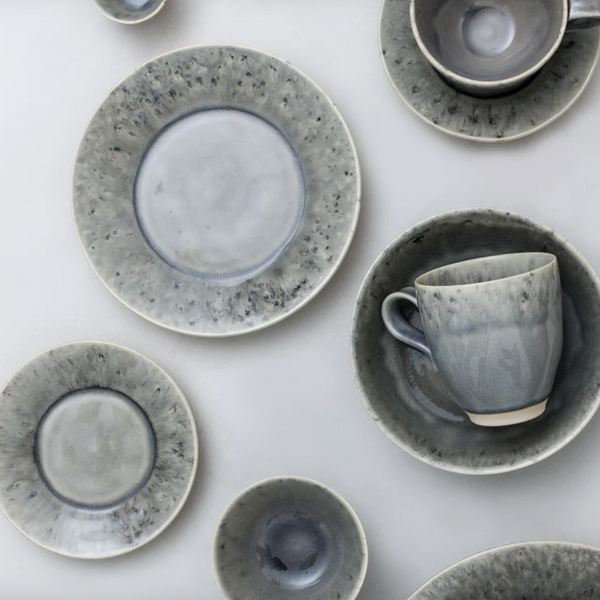 Costa Nova’s Madeira Grey Dinnerware features stoneware plates, bowls, and cups in mottled grey and white finishes, artistically arranged on a light surface.