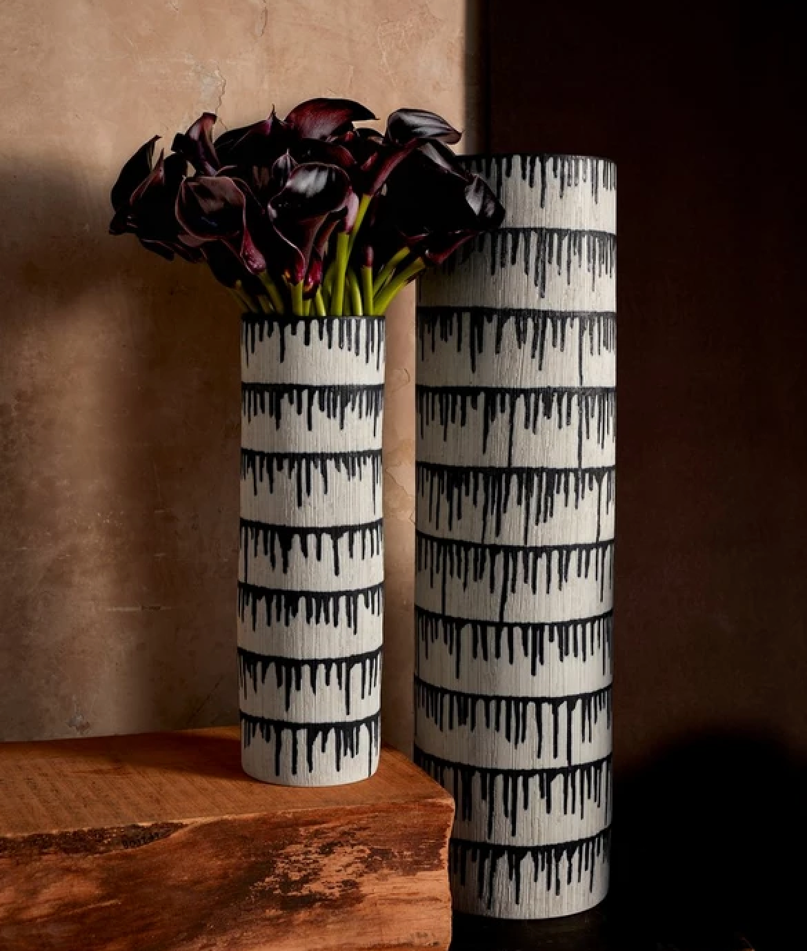 Two tall, cylindrical Tokasu Vases by L'Objet, featuring a pattern inspired by Japanese indigo dyeing, are displayed on wood. One holds dark purple calla lilies while the other stands empty against a textured neutral background.