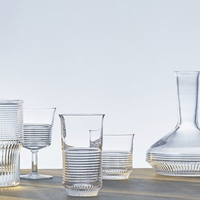 Cadence Barware by Saint-Louis/Hermes, featuring a crystal tumbler and wide-bottomed carafe with ribbed or fluted textures, is displayed on a light surface. The glassware sits against a minimal, softly lit background.