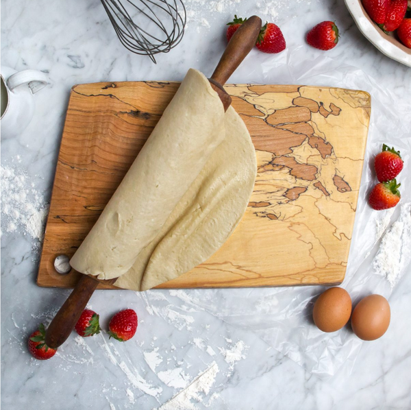A rolling pin with dough sits on the Peterman Spalted Maple Rectangle Cutting Board 18". Fresh strawberries, two eggs, and baking utensils are scattered on a flour-dusted marble surface.