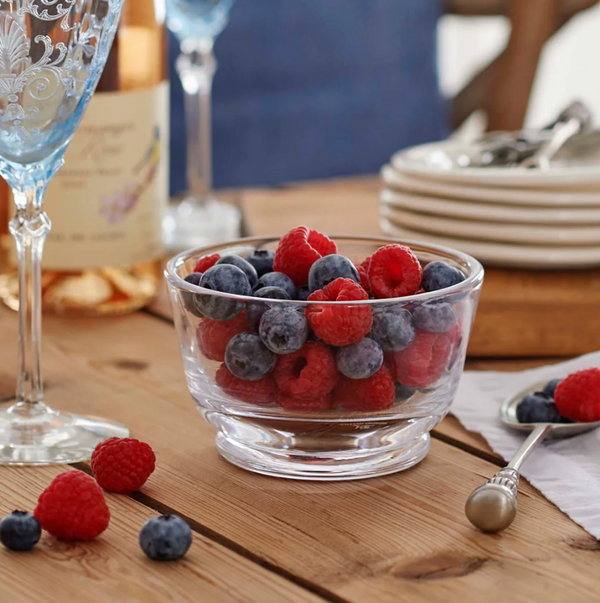 The William Yeoward Corinne Fruit / Nut Bowl, filled with fresh raspberries and blueberries, sits on a wooden table beside an elegant wine glass, stacked plates, a spoon, and scattered berries.