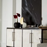 A modern sideboard with gold handles is adorned with a vase of red flowers, the Arteriors Hiroshi Sculpture—a round gold object—elegant dishes, and topped by a striking black-and-white abstract painting evocative of cubist art.