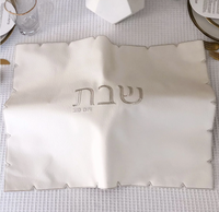 The BT-Shalom White Embroidered Leatherette Challah Cover - Silver features a textured, laser-cut edge and central Hebrew text, displayed on a white tablecloth with gold-rimmed utensils and glassware set for a meal.