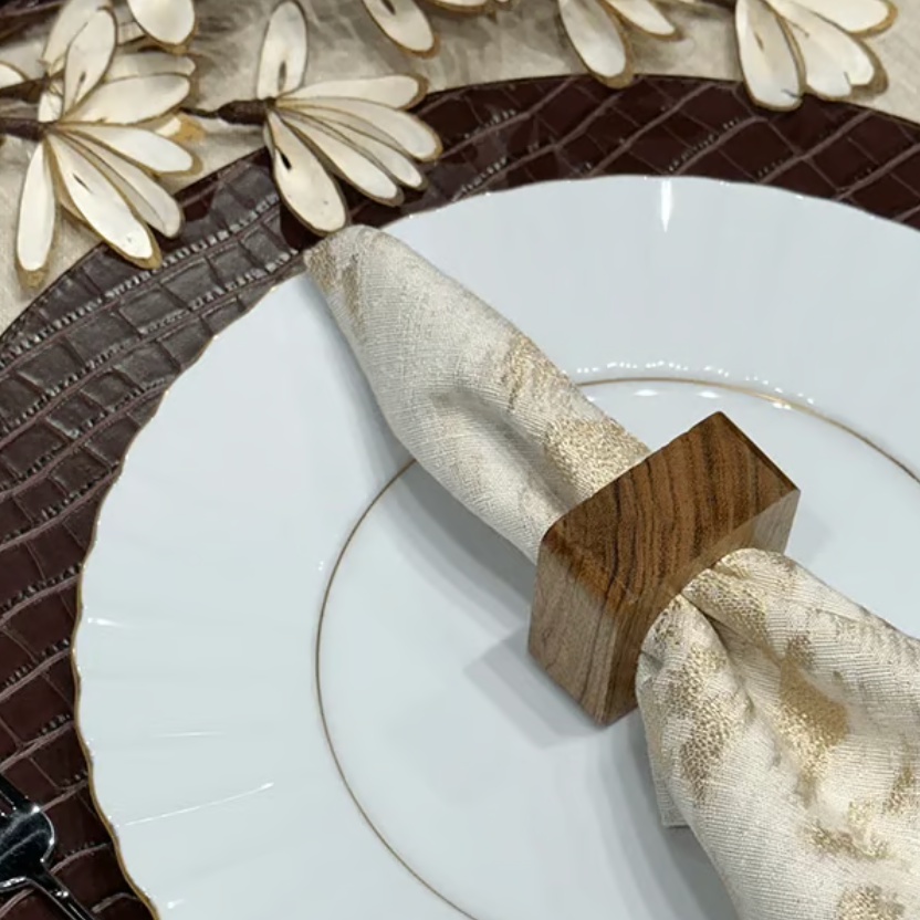 A white plate with a beige patterned napkin is held by the Von Gern Home Mocha Napkin Ring Brown (Set of 4) and placed on a brown textured placemat, adorned with cream and gold leaf accents.