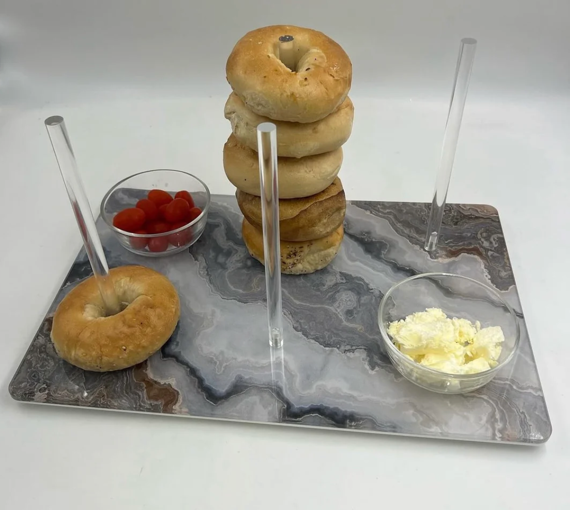 The Eye Candy Continental Bagel Board in Grey Marble Slice holds four clear rods—one stacked with bagels, one with a single bagel, and two empty. Small bowls of cherry tomatoes and cream cheese complete this elegant display.