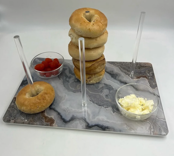 The Eye Candy Continental Bagel Board in Grey Marble Slice holds four clear rods—one stacked with bagels, one with a single bagel, and two empty. Small bowls of cherry tomatoes and cream cheese complete this elegant display.