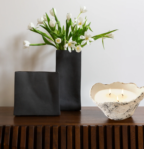 A dark rectangular handmade vessel with white tulips and a matching square object rests on wood beside a white textured bowl displaying the glowing Dumae Gemma Candle - Sierra Mista - 10", with two flames.