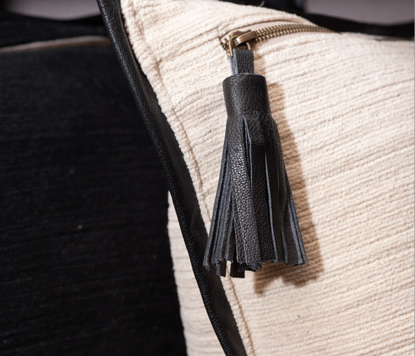 A close-up of the Threads Soft Goods Leather Frame Pillow Ivory showcases cream fabric, black piping, and a black leather tassel zipper pull.