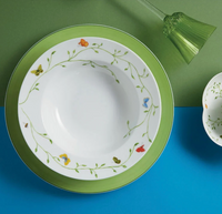 The Histoire Naturelle Dinnerware by Raynaud features a white bowl with green rim, vine, butterfly, and rare bird designs on a matching plate, set beside a green glass and patterned dish on a blue-green background for an elevated dining experience.