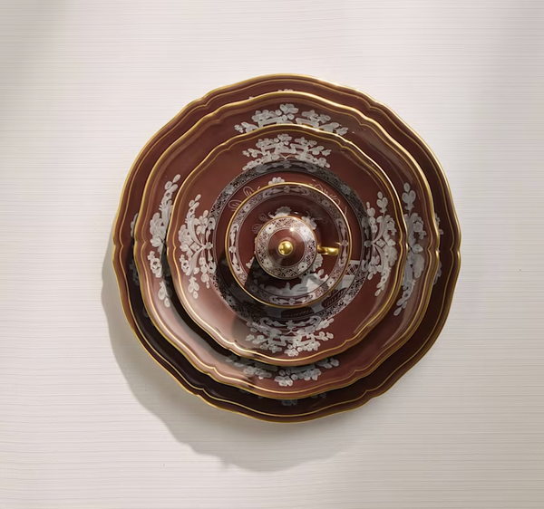 Concentrically arranged on a light surface, Ginori 1735’s Oriente Italiano Castagna brown and gold plates with a lidded cup display elegant white floral designs, showcasing the Italian-made tableware set from a top view.