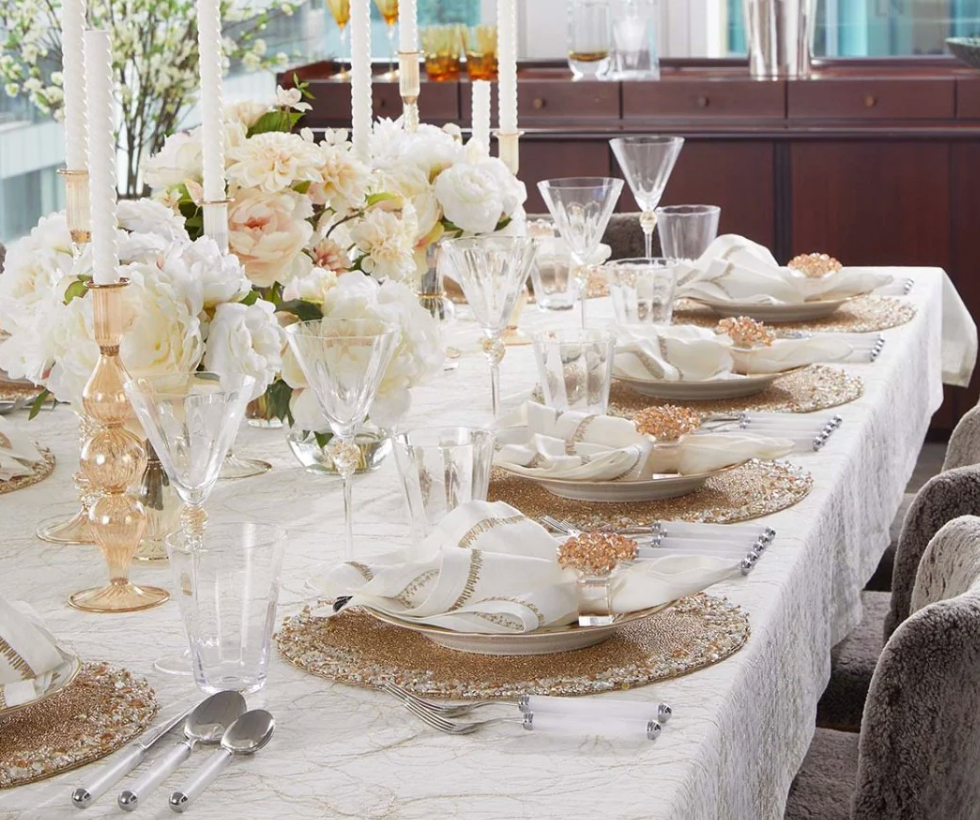 Create a luxurious, festive table with white and gold tableware, crystal glasses, tall candles, a white floral centerpiece, and folded napkins adorned with the Kim Seybert Aster Napkin Ring Set of 4.