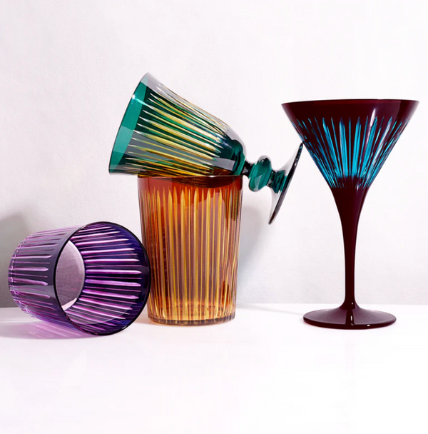 The Prism Wine Glass Set of 4 by L'Objet features colorful glasses in purple, amber, green, and blue with vertical ribbed patterns, arranged playfully on a white surface.