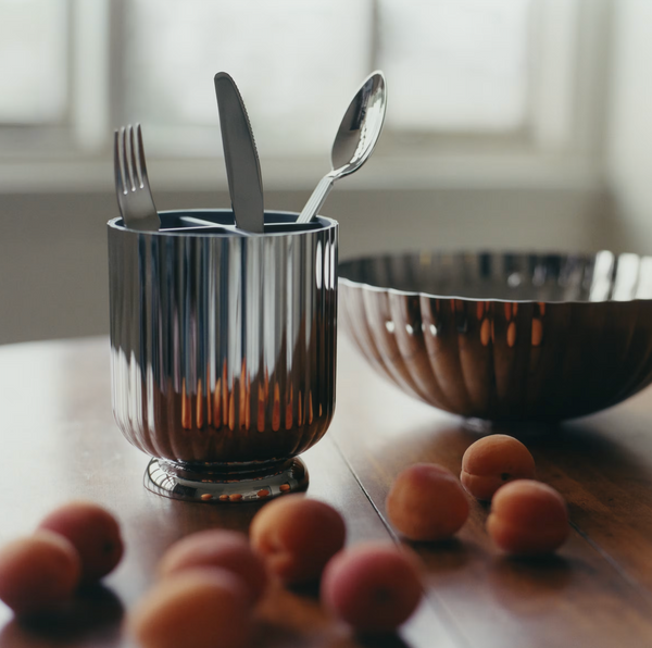 A Georg Jensen Bernadotte Cutlery Holder cradles a fork, knife, and spoon on a wooden table with apricots and a matching stainless steel bowl in soft natural light.