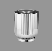 The Georg Jensen Bernadotte Cutlery Holder is a shiny stainless steel piece with vertical grooves, a round base, and an internal cross-shaped divider, displayed against a plain gray background.