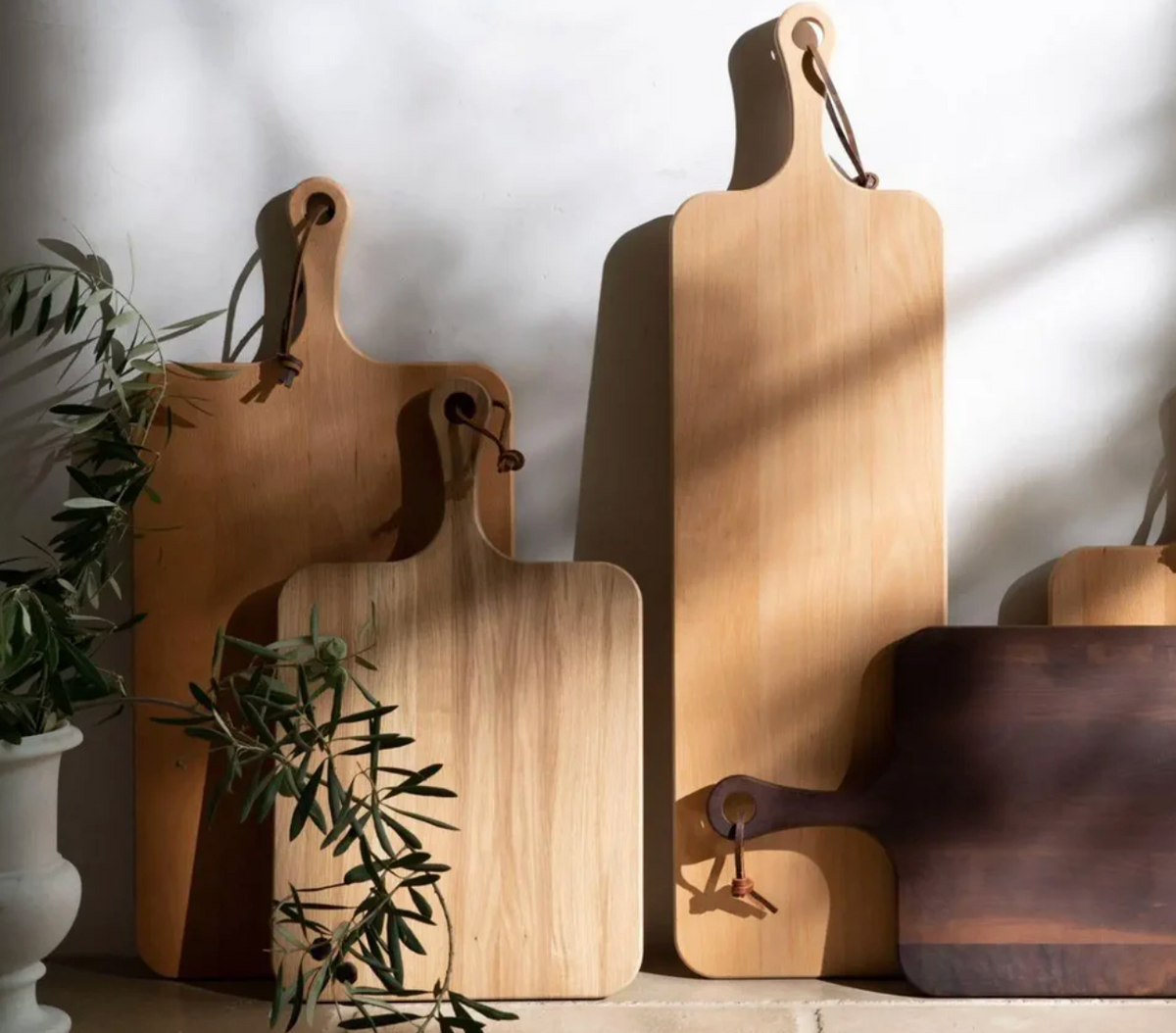 Several wooden cutting boards, including the Edmund Narrow Oak Serving Board by Blue Pheasant, stand upright against a white wall—some with leather straps. Sunlight casts shadows as green leaves frame the serving board in the foreground.