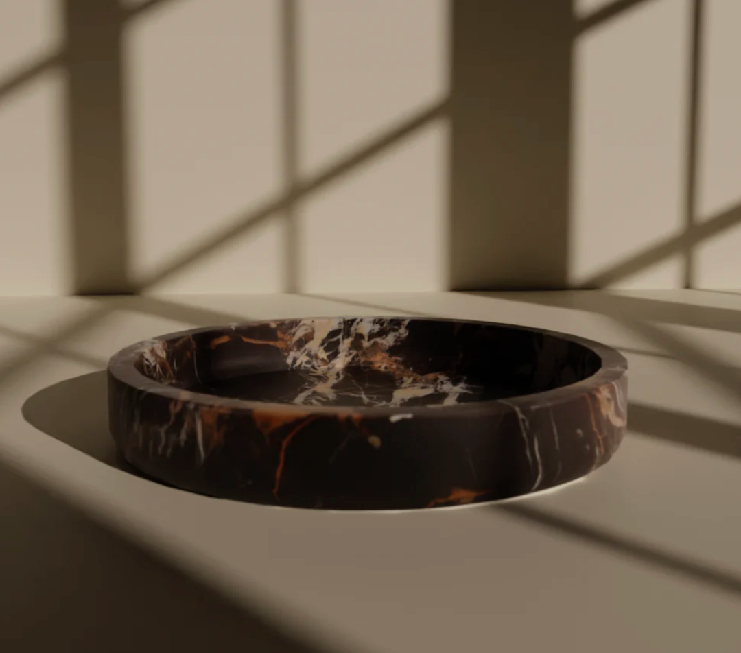 A round, shallow Flat Honed Marble Bowl by Designs by Marble Crafter in stunning black and gold rests on a sleek surface, sunlight streaming through window panes casting geometric shadows around it. This statement piece serves as an elegant focal point for the space.