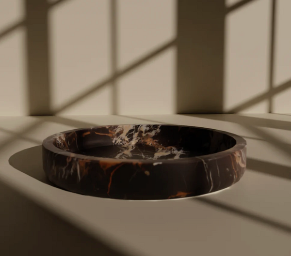 A round, shallow Flat Honed Marble Bowl by Designs by Marble Crafter in stunning black and gold rests on a sleek surface, sunlight streaming through window panes casting geometric shadows around it. This statement piece serves as an elegant focal point for the space.