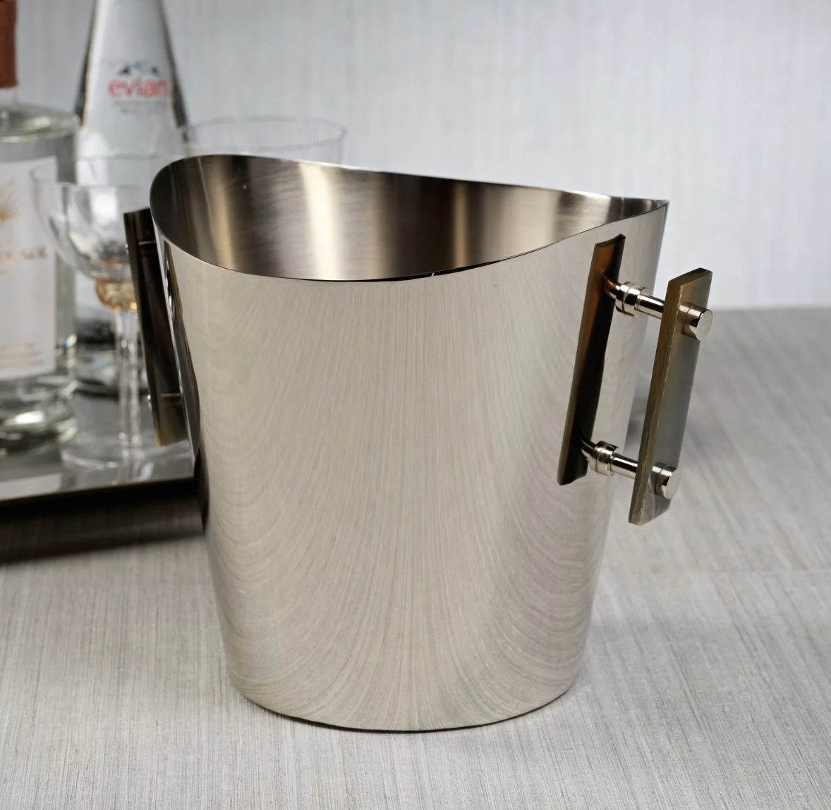The Zodax Belmont Wine Cooler / Ice Bucket - Small, featuring two rectangular side handles, sits on a light surface with a tray of bottles and glasses subtly blurred in the background.