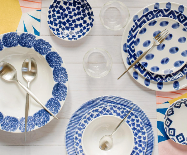 The Vietri Santorini Assorted Cocktail Plates Set of 4 features blue and white patterns, paired with metal utensils and glass tumblers on a white wood table with colorful geometric napkins—dishwasher safe for easy cleanup.