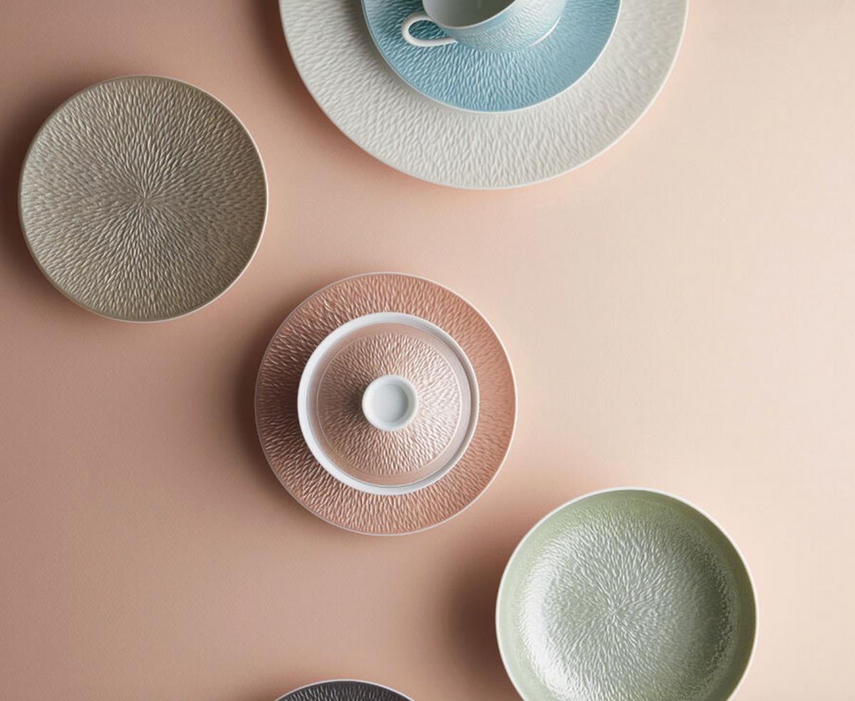 Raynaud’s Mineral Irise Dinnerware in Copper features luxurious porcelain plates, bowls, and a cup with saucer, elegantly arranged on a light peach background to showcase their refined beauty.