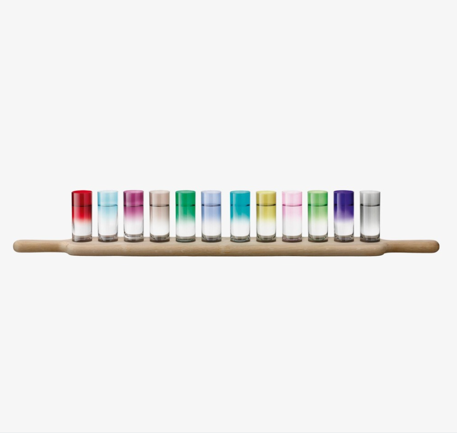 Twelve vibrant ombre shot glasses from the LSA Paddle Grand Shot Set are arranged on an oak paddle, showcasing shades of red, pink, blue, green, yellow, and purple—an eye-catching party set.