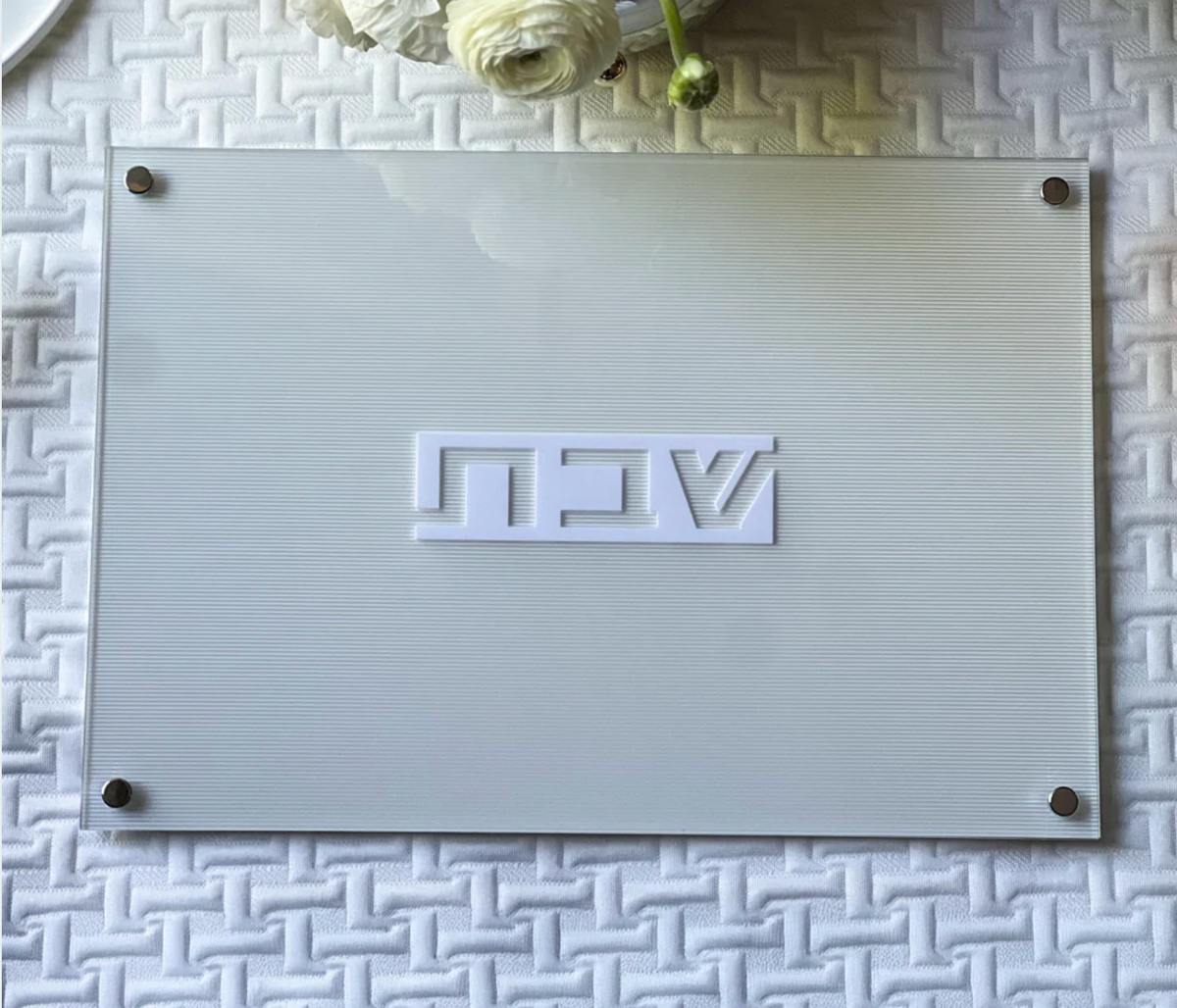 The BT-Shalom Lucite Corrugated Challah Board features a textured white surface with raised white Hebrew letters and four metal corner fasteners—a sleek, modern centerpiece. White flowers and a plate are partially visible in the upper left.