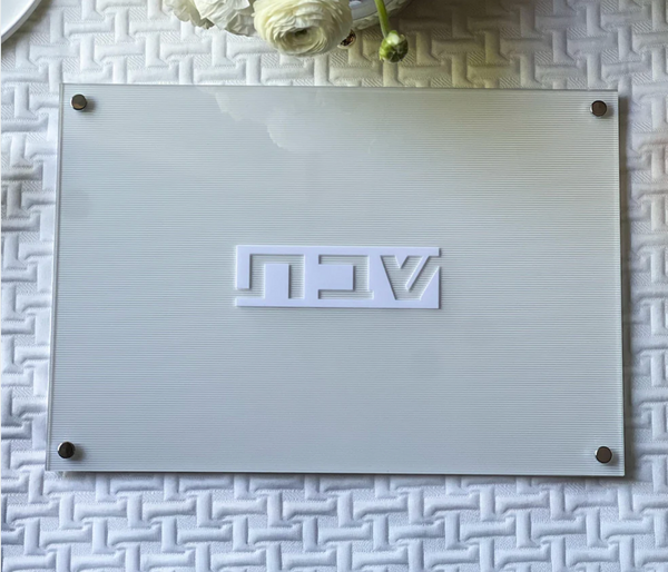 The BT-Shalom Lucite Corrugated Challah Board features a textured white surface with raised white Hebrew letters and four metal corner fasteners—a sleek, modern centerpiece. White flowers and a plate are partially visible in the upper left.
