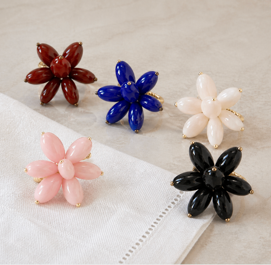 The Mod Stone Flower Napkin Ring Set of 4 by Deborah Rhodes, in vibrant petal-inspired red, blue, white, pink, and black with gold accents, is arranged on a light surface beside a white napkin.