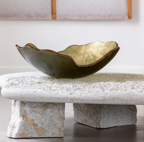 The Gardeco Mapa Gold & Bronze Trim Bowl, featuring a wavy edge and radiant finish, is displayed on a textured stone coffee table with block-style legs in a modern minimalist room.