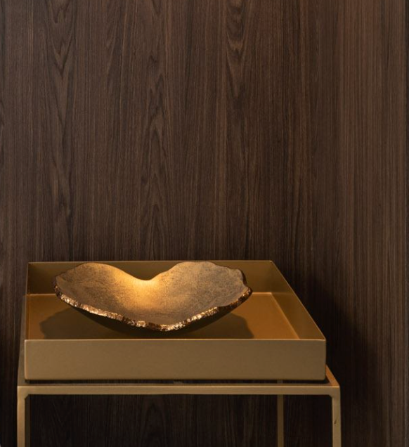 The Gardeco Mapa Gold & Bronze Trim Bowl, a metallic heart-shaped artisan piece, sits on a golden tray atop a stand before a dark wood-paneled wall, its rich textures and tones enhanced by warm lighting.