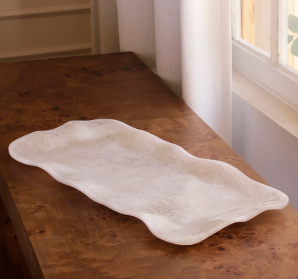 The Beatriz Ball Resin Alabaster Long Rectangle Tray - White Swirl sits on a wooden table near a window with cream-colored curtains.