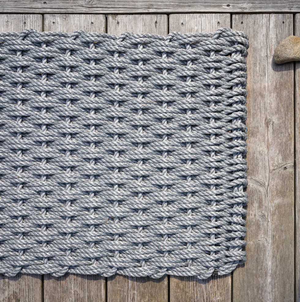 The Rope Co Coastal Woven Outdoor Floormat in Weathered Gray (24 x 38) rests on a weathered wooden deck, with the edge of the mat visible near a round stone in the upper right corner.