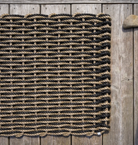 The Rope Co's Coastal Woven Outdoor Floormat in Sand & Charcoal (24 x 38) sits on a wooden deck, highlighting its durability with a large stone placed on the top right corner.