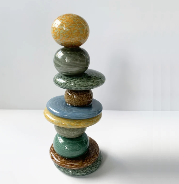 Axiom’s Pohacu Sculpture features ten stacked, glossy, colorful ceramic stones—spheres and discs with rounded shapes—balanced vertically on a white surface against a plain light background, echoing the style of Robert Madvin’s stacked stone artworks.