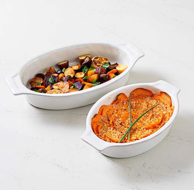Two Juliska Puro Oval Baker Whitewash 16″ stoneware bakers: one filled with roasted mixed vegetables, the other with sweet potato casserole topped with breadcrumbs and garnished with chives, both displayed on a light surface for effortless oven-to-table serving.