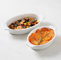Two Juliska Puro Oval Baker Whitewash 16″ stoneware bakers: one filled with roasted mixed vegetables, the other with sweet potato casserole topped with breadcrumbs and garnished with chives, both displayed on a light surface for effortless oven-to-table serving.